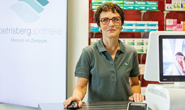 Apothekerin der Petrisberg Apotheke in Trier steht hinter dem Tresen am Pc. Hinter Ihr sieht man das Medikamentenregal, dass unterstreicht die Fachkompetenz und technische Ausstattung der Apotheke.