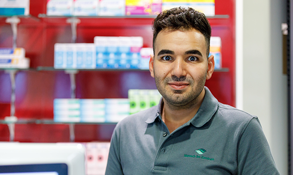 Der Apotheker und Inhaber der Petrisberg Apotheke steht an der Kasse vor dem Medikamentenregal und blickt in die Kamera.
